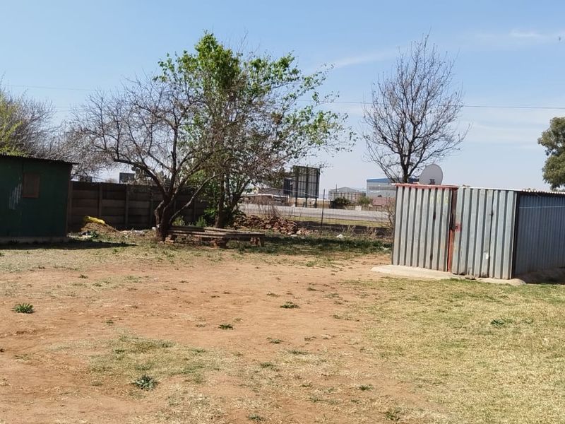 A large, unmaintained yard featuring two corrugated metal outbuildings and a precast concrete boundary wall.