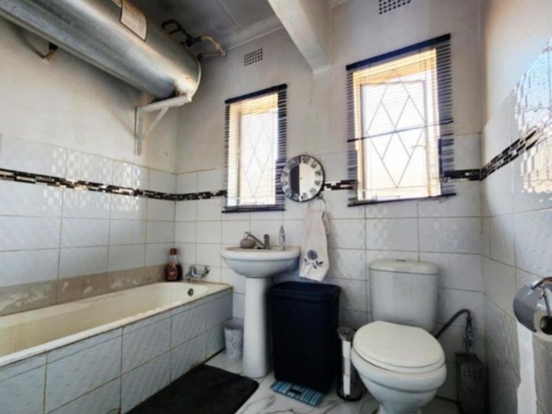 A functional bathroom featuring a bathtub, pedestal basin, and toilet, with wall tiling and an exposed electric geyser unit.