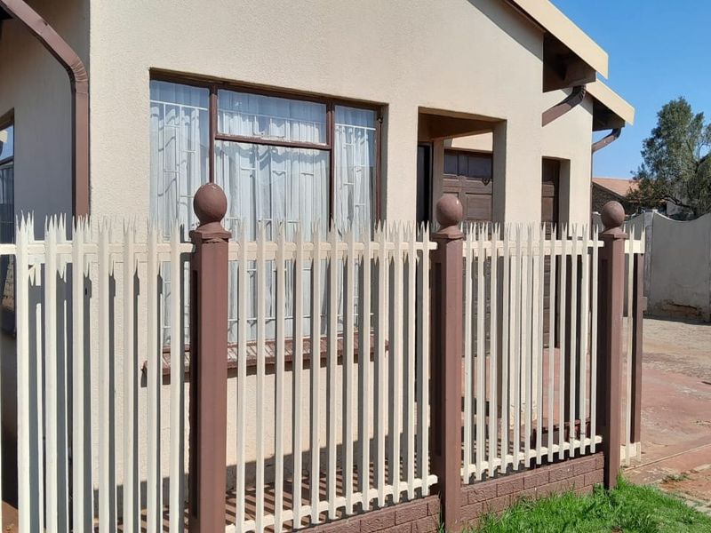 Front elevation of a residential house featuring a secure metal fence, paved driveway, and standard window security bars.