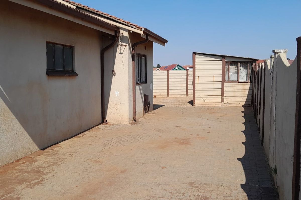 Rear exterior view showing a paved driveway, boundary wall, and the side elevation of the house with visible plaster cracking.