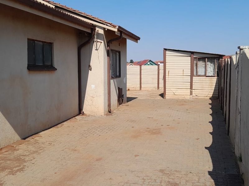 Rear exterior view showing a paved driveway, boundary wall, and the side elevation of the house with visible plaster cracking.