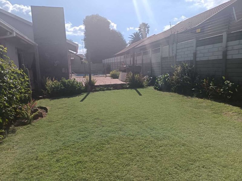 A private rear garden featuring a well-maintained lawn, paved patio area with a built-in braai, and a swimming pool enclosed by brick boundary walls.