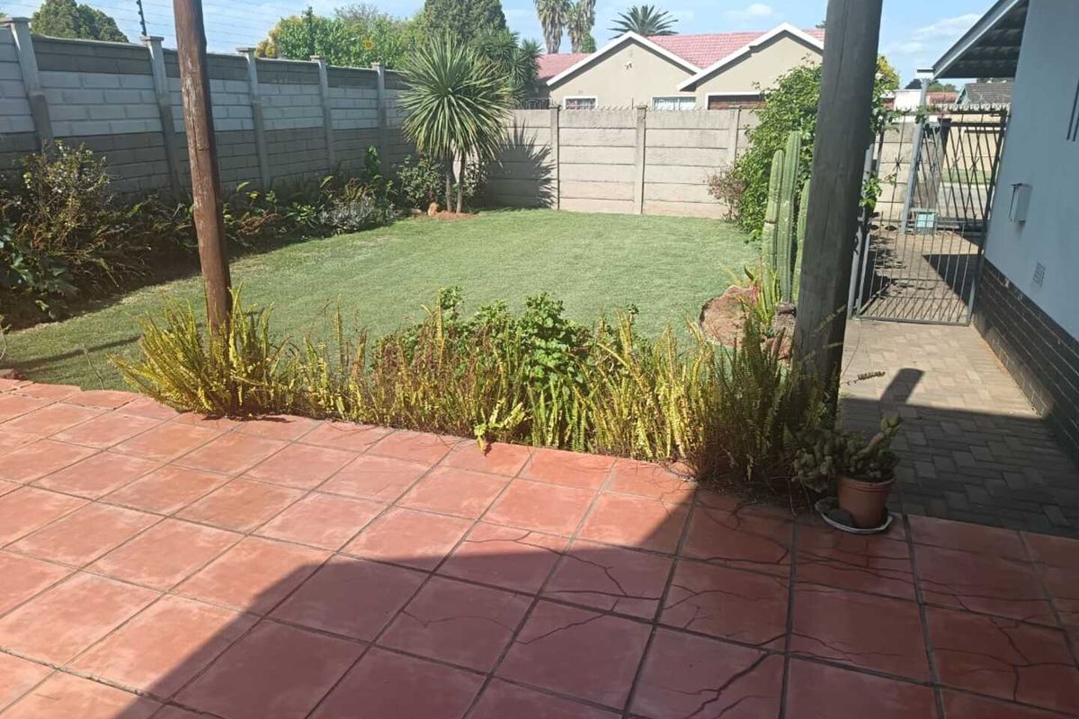 A rear garden area featuring a tiled patio with visible cracking, a well-maintained lawn, and secure perimeter walling.