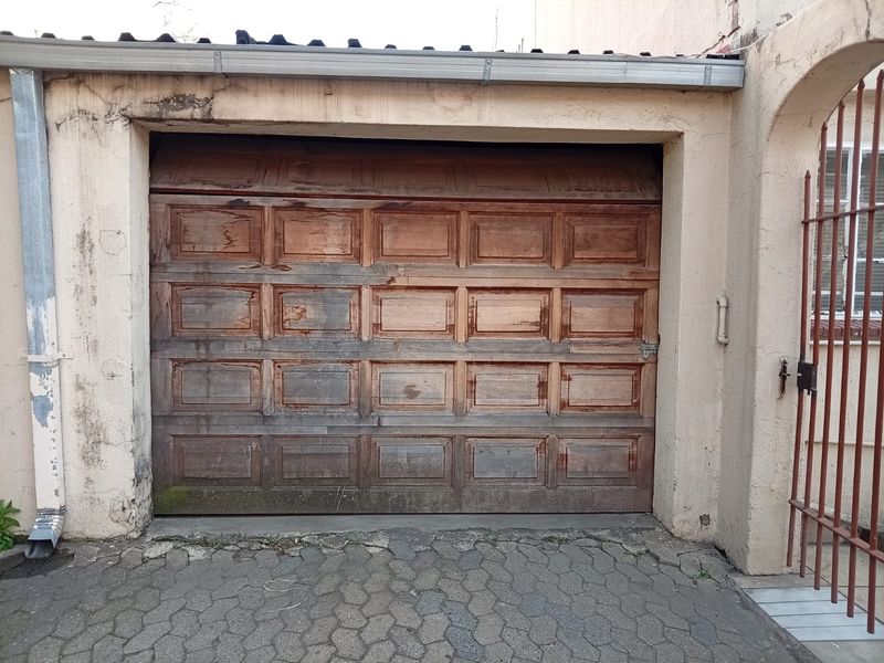 Exterior view of a single garage featuring a weathered wooden sectional door, adjacent metal security gate, and paved driveway area.