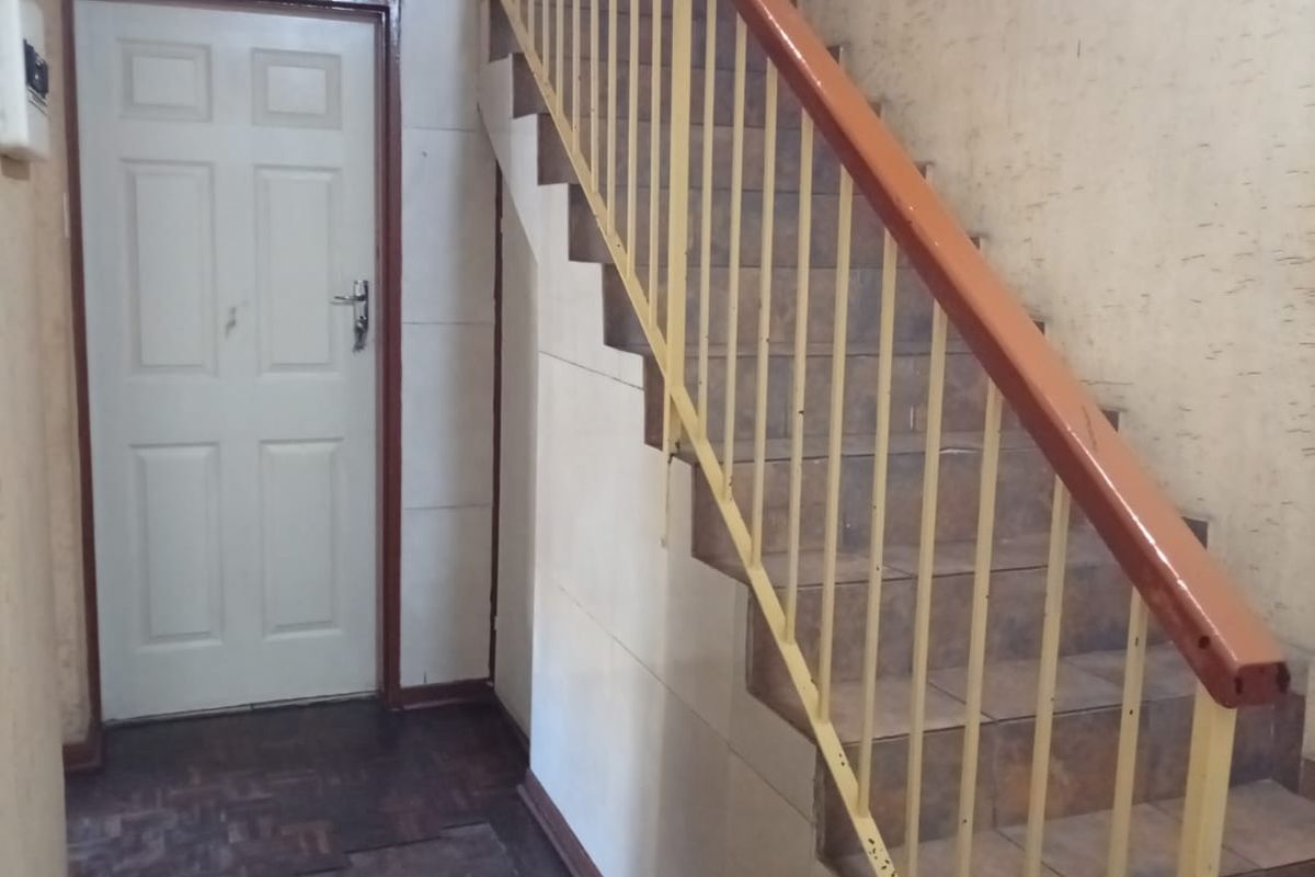 A hallway featuring a staircase with a metal balustrade, tiled steps, and parquet flooring, showing signs of wear and requiring cosmetic updates.