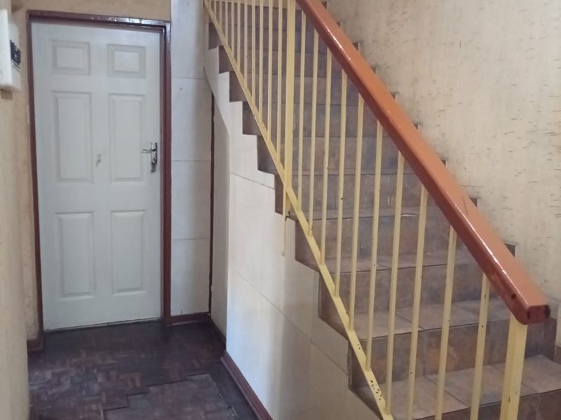 A hallway featuring a staircase with a metal balustrade, tiled steps, and parquet flooring, showing signs of wear and requiring cosmetic updates.