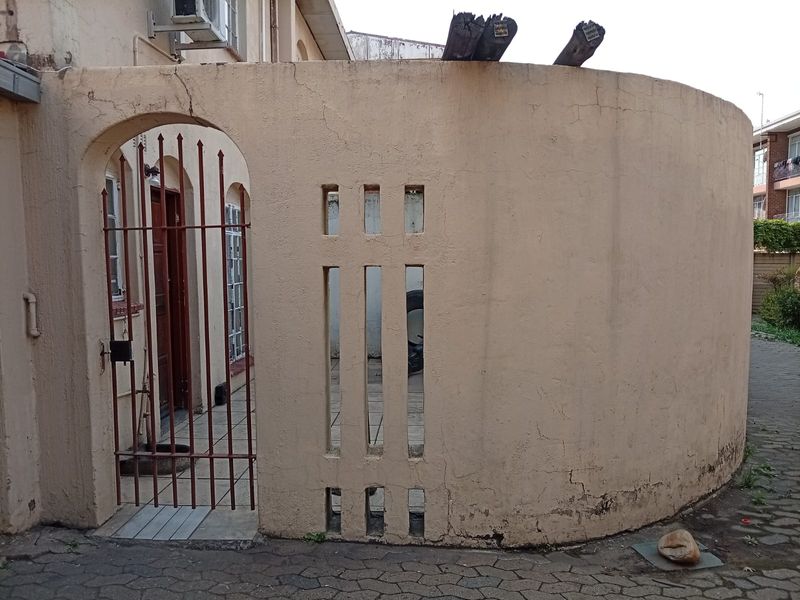 Exterior view of an apartment entrance featuring a curved masonry wall, wrought iron gate, and tiled entryway with visible signs of weathering.