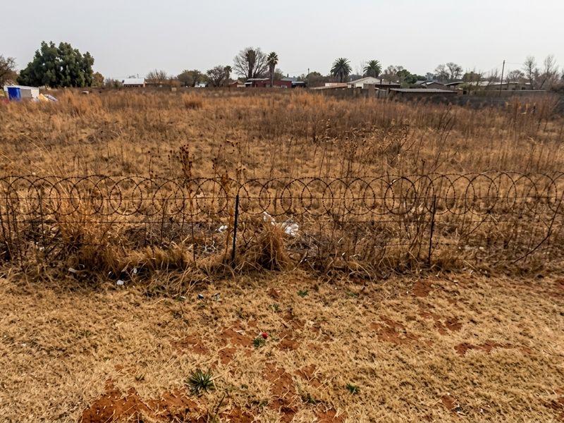 A vacant residential plot featuring dry, overgrown grass and secured by a wire mesh fence topped with razor wire.
