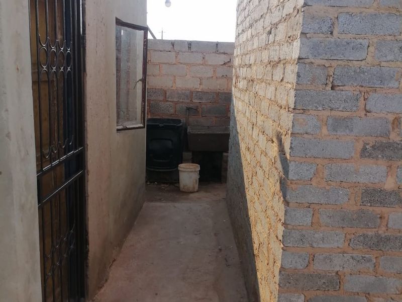 A narrow rear exterior passage featuring a concrete pathway, security gate, and an external utility sink area with unfinished brick boundary walls.