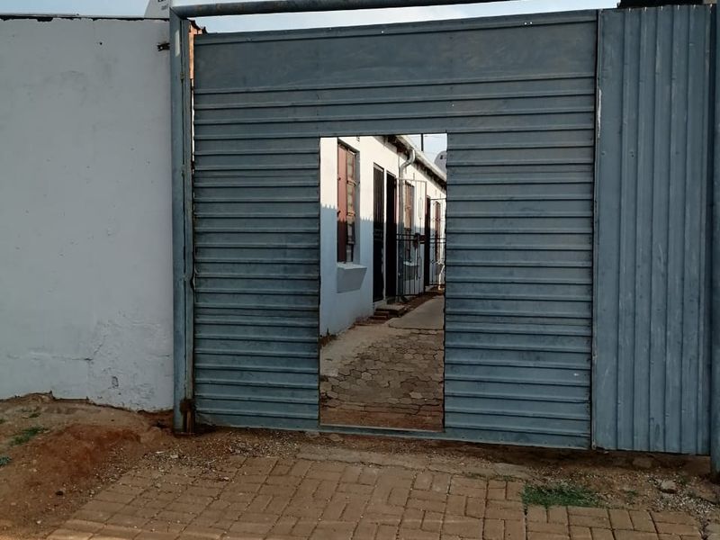 Front entrance view featuring a corrugated metal gate, paved driveway, and boundary wall leading to the property exterior.