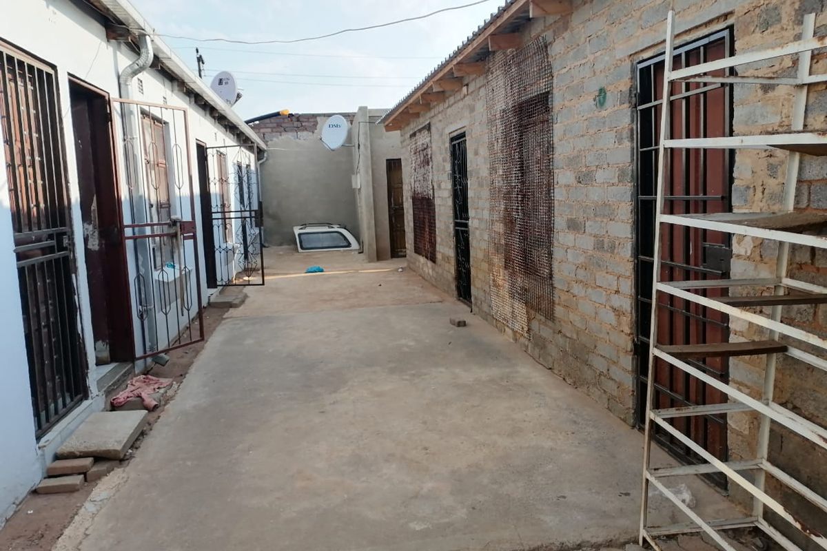 A narrow exterior rear courtyard area featuring concrete paving, exposed brick walls, and multiple security-gated entrances to the apartment units.