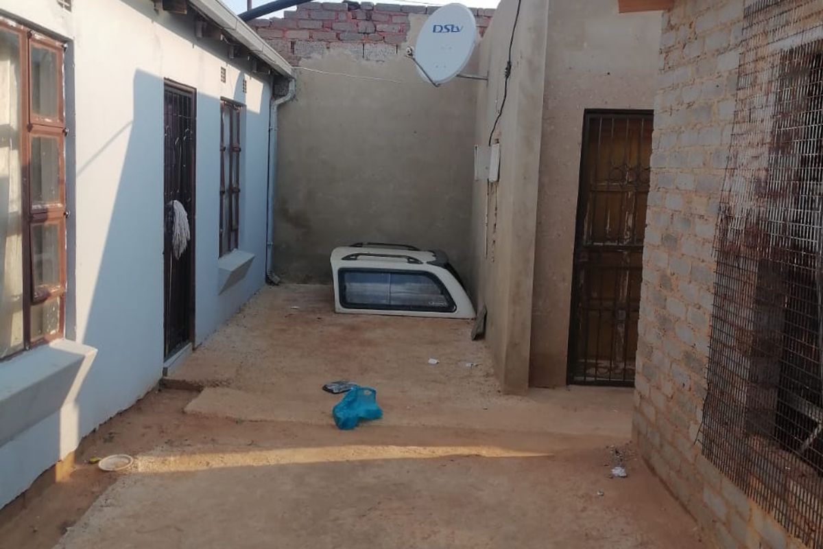 Rear exterior area featuring exposed brickwork, security gates, and satellite dish installation, with an unpaved ground surface between two building structures.
