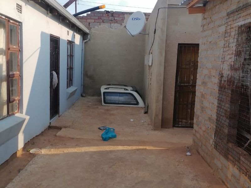 Rear exterior area featuring exposed brickwork, security gates, and satellite dish installation, with an unpaved ground surface between two building structures.
