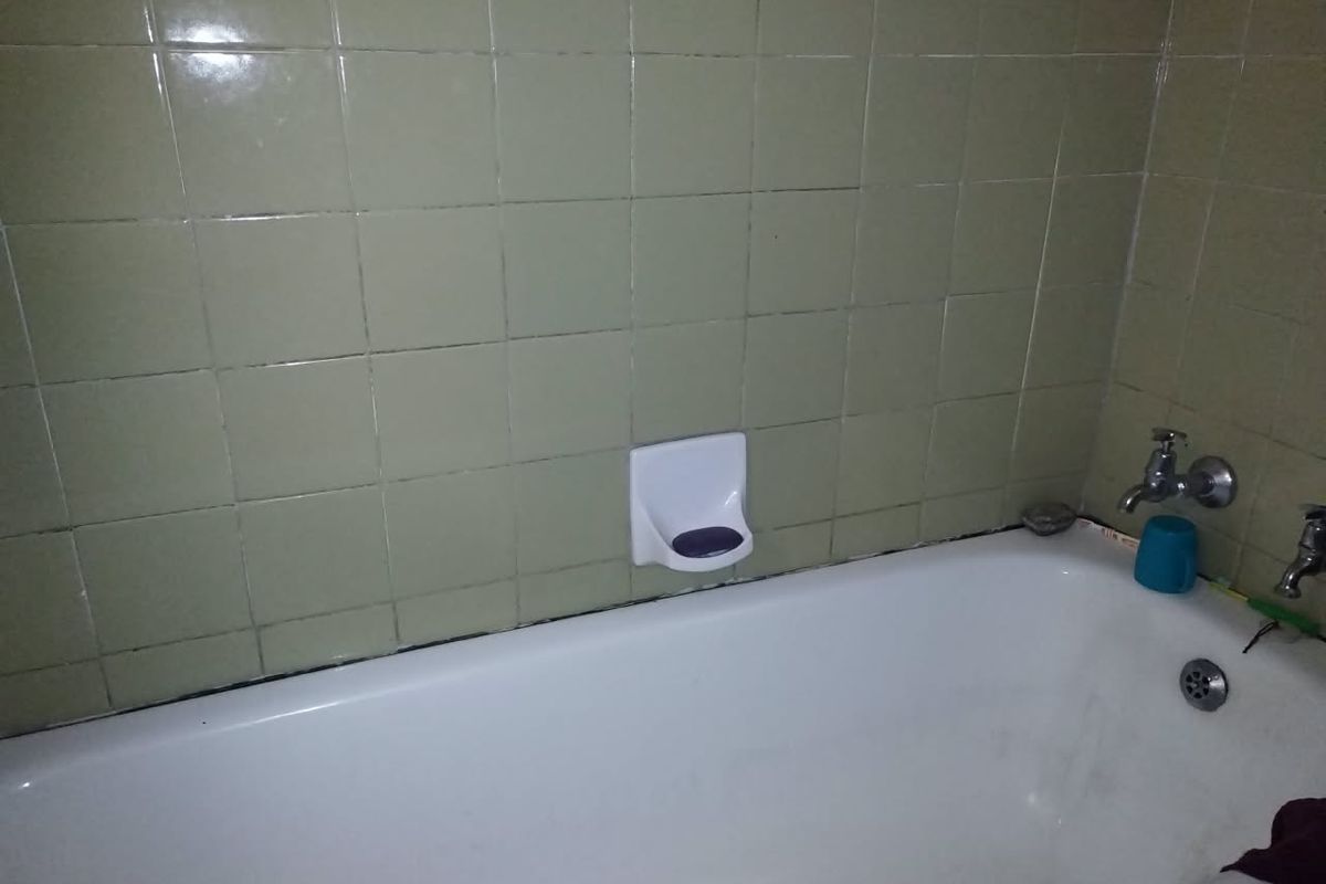 A dated bathroom featuring a standard bathtub with wall-mounted fixtures and olive-toned ceramic wall tiling.