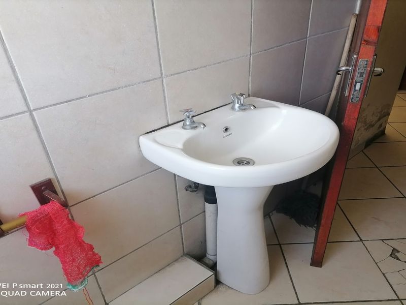 A functional bathroom space featuring a pedestal basin and tiled surfaces, currently showing signs of wear including cracked floor tiles and damaged door trim.