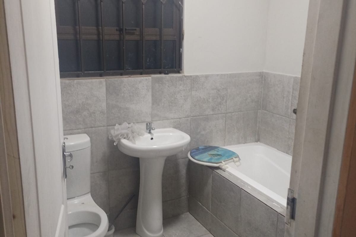 A functional bathroom featuring a pedestal basin, toilet, and bathtub with neutral grey wall and floor tiling and a security-fitted window.