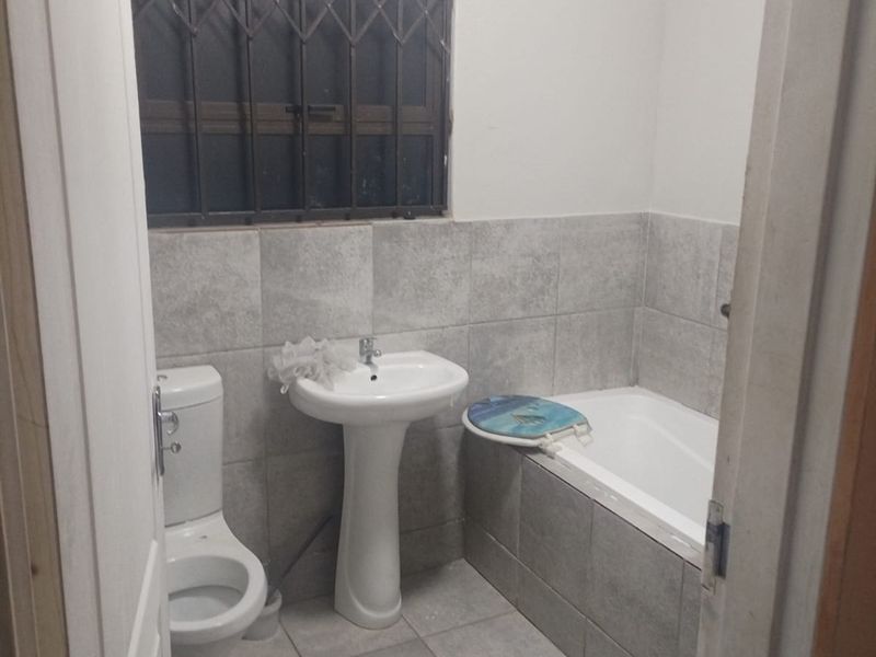 A functional bathroom featuring a pedestal basin, toilet, and bathtub with neutral grey wall and floor tiling and a security-fitted window.