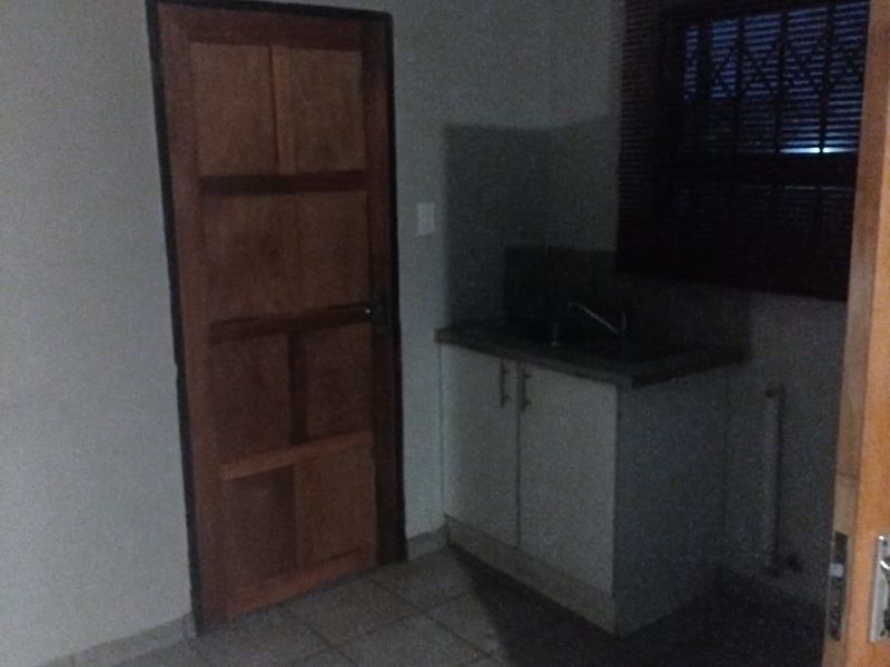 A small, empty scullery or utility area featuring tiled floors, a built-in sink cabinet, and a window with security bars.