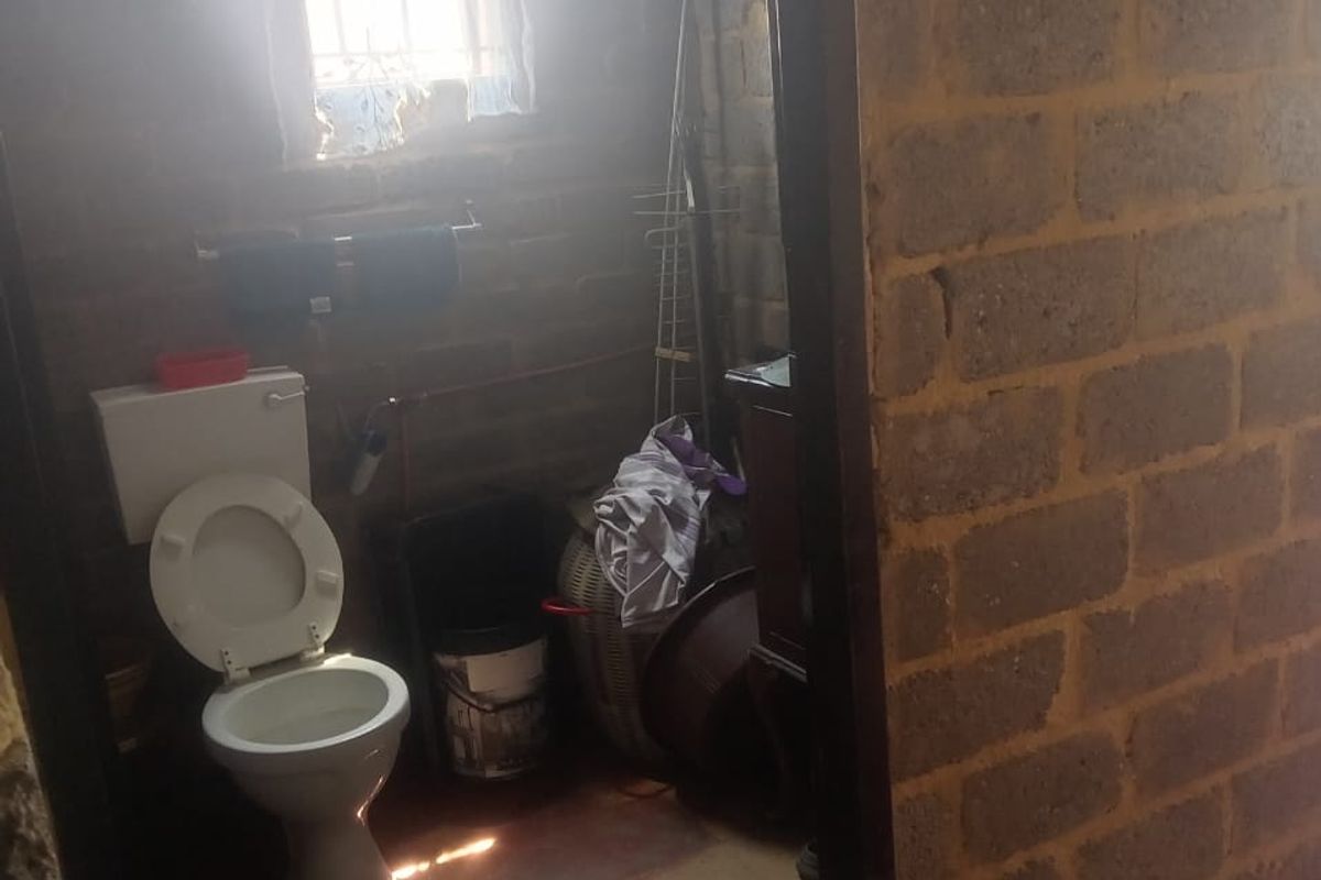 A small bathroom featuring exposed brick walls, a standard toilet, and a window with security bars, requiring significant renovation and finishing.
