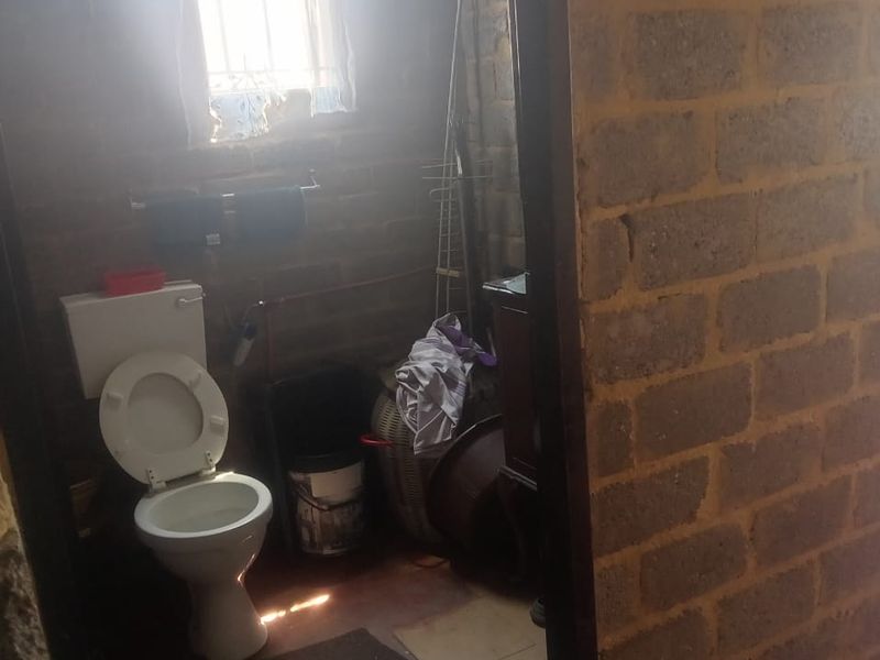 A small bathroom featuring exposed brick walls, a standard toilet, and a window with security bars, requiring significant renovation and finishing.