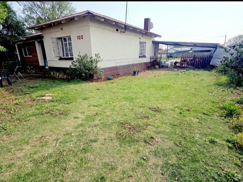Rear view of a single-story house featuring a corrugated metal roof, brick chimney, and an attached carport, set within a spacious backyard.