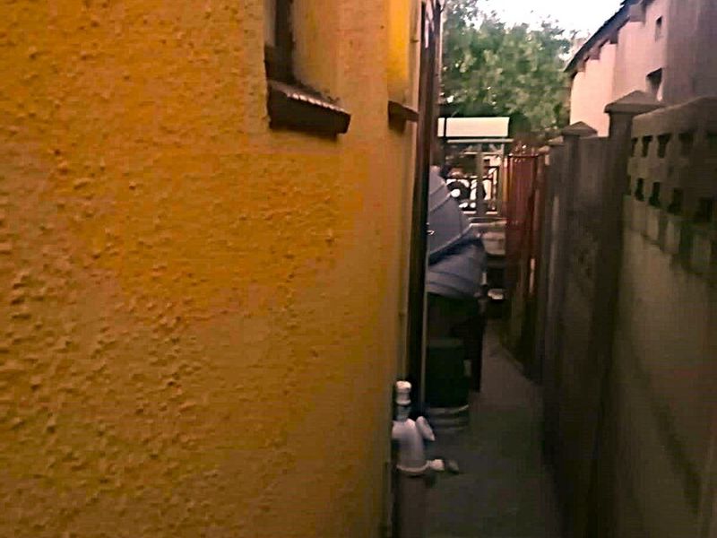 A narrow side passage between the main house structure and a concrete boundary wall, showing weathered exterior finishes.