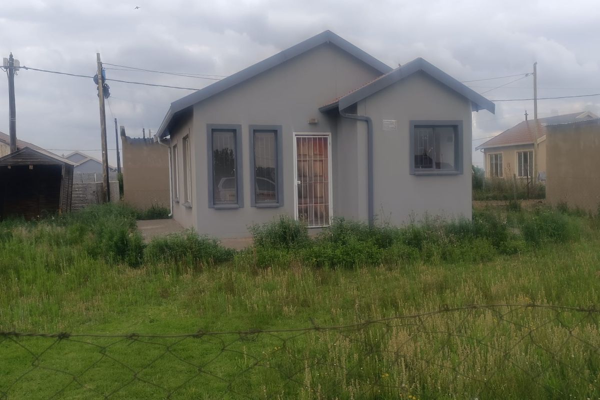 A detached single-story house featuring a gabled roof, grey exterior finish, and security gate, situated on a large, grass-covered plot.