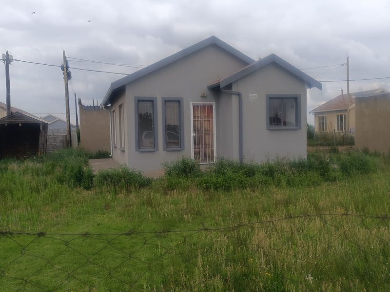 A detached single-story house featuring a gabled roof, grey exterior finish, and security gate, situated on a large, grass-covered plot.