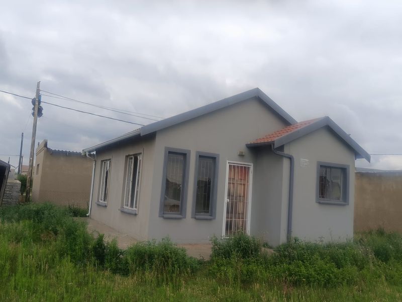 A detached single-story house featuring a grey facade, tiled roof, and security bars on windows, set on an unpaved plot with overgrown grass.