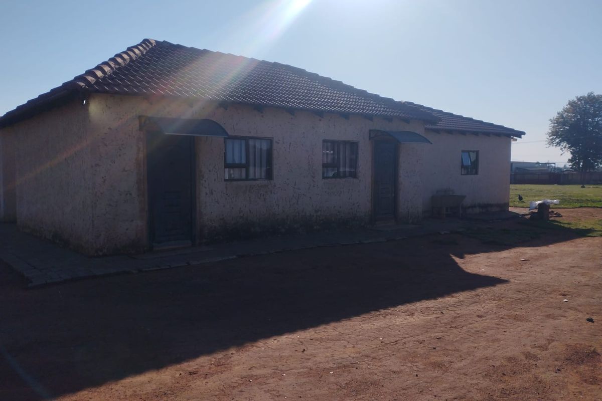 A single-story residential building featuring a tiled roof and plastered exterior walls, situated on a large plot with a dirt driveway.