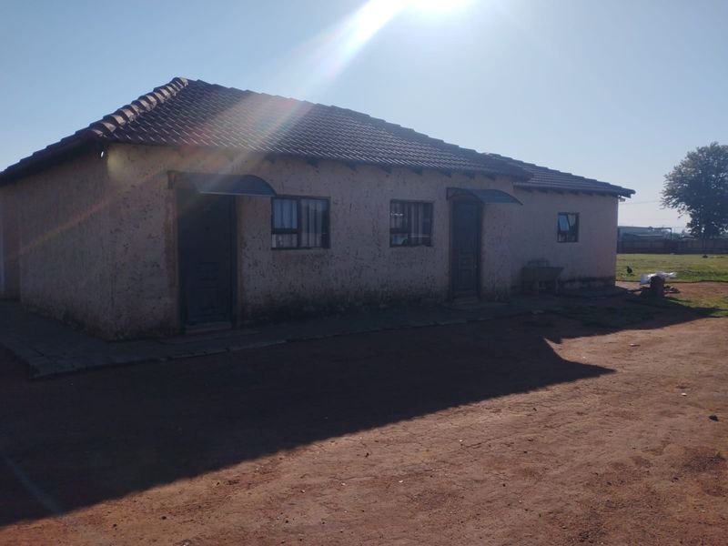 A single-story residential building featuring a tiled roof and plastered exterior walls, situated on a large plot with a dirt driveway.