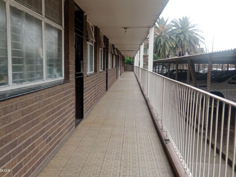 An exterior covered walkway featuring brick walls, tiled flooring, and a metal balustrade overlooking the parking area.