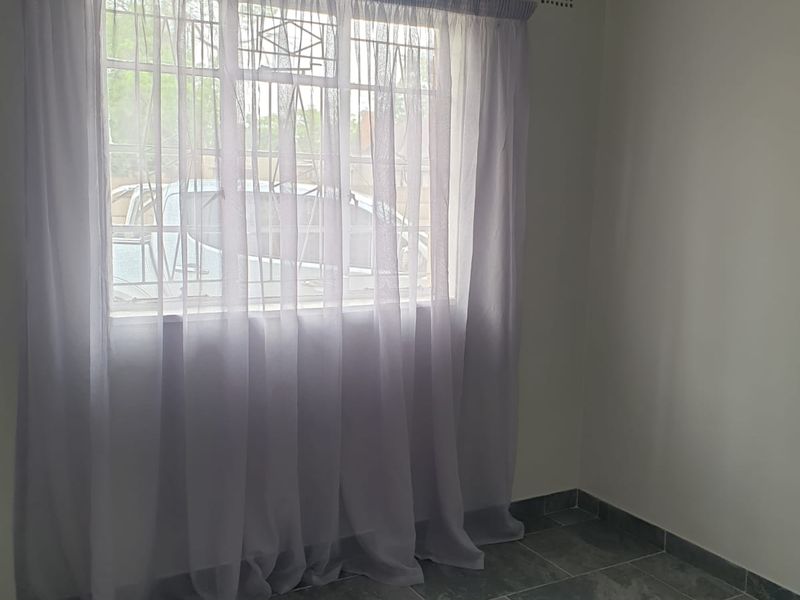 A clean, empty bedroom featuring neutral tiled flooring, a window with security bars, and a ventilation grille.