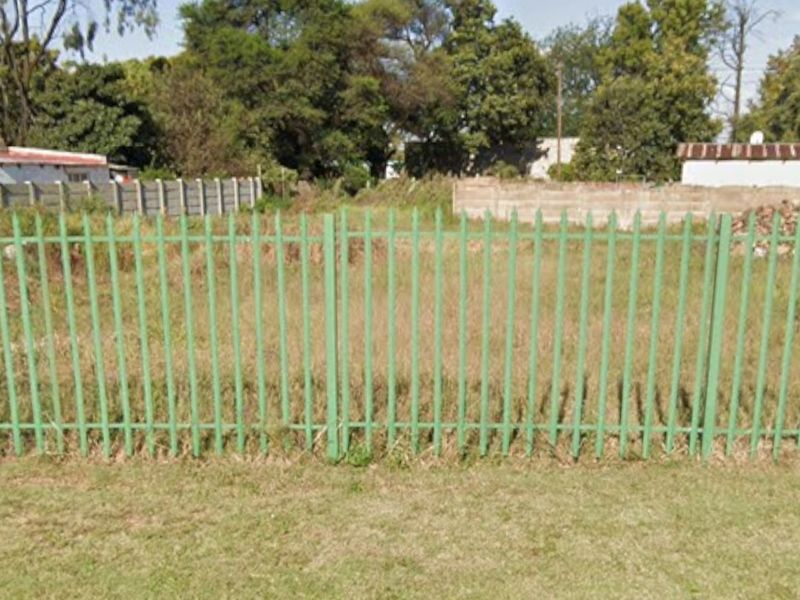 A vacant residential plot enclosed by metal palisade fencing and concrete boundary walls, featuring overgrown grass and established trees in the background.