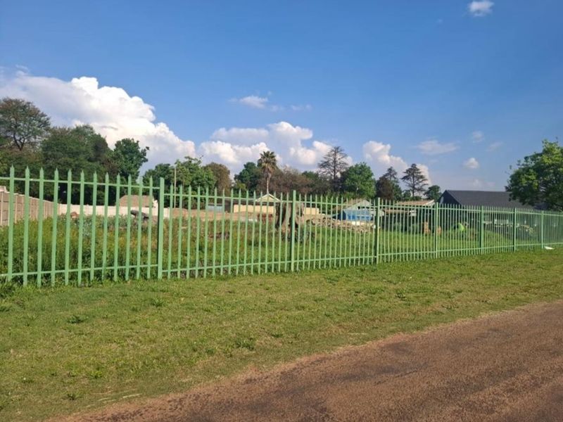 A view of a vacant residential plot secured with green metal palisade fencing, bordering a dirt road.