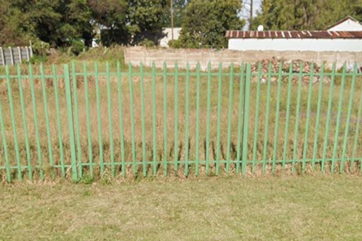 A vacant residential plot featuring green metal palisade fencing and a concrete boundary wall, with overgrown grass and established trees in the background.