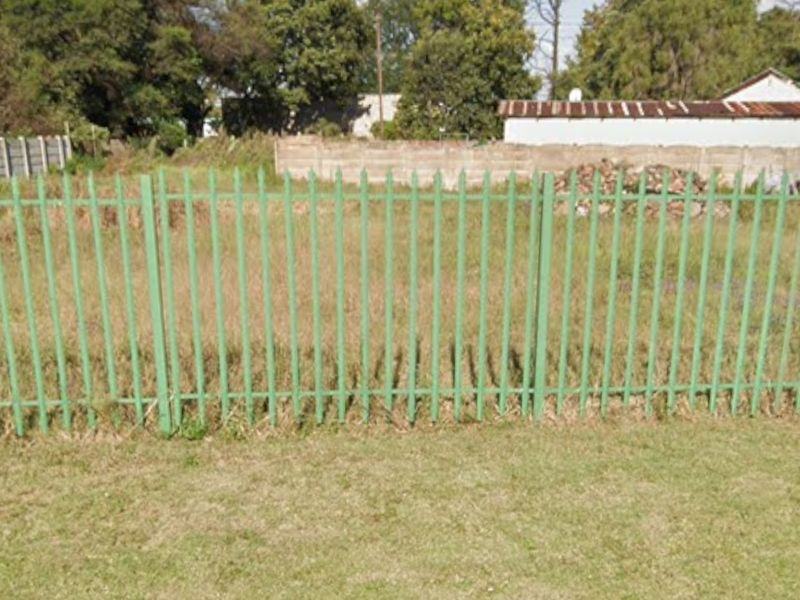 A vacant residential plot featuring green metal palisade fencing and a concrete boundary wall, with overgrown grass and established trees in the background.