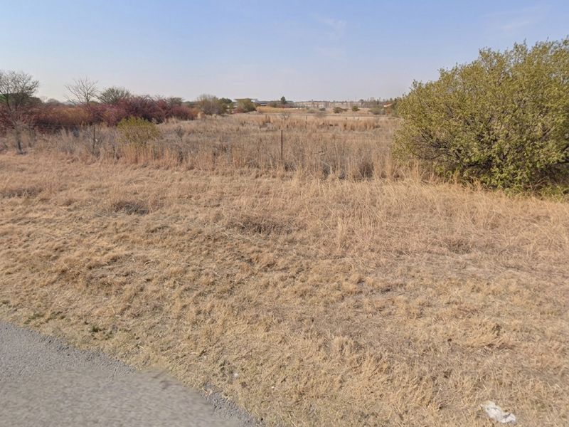 A view of a vacant residential plot featuring dry grass, natural vegetation, and perimeter wire fencing along the roadside.