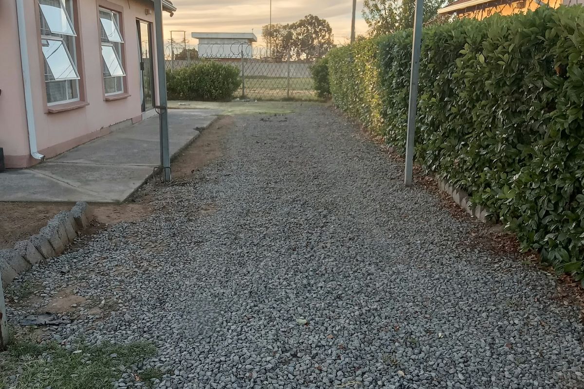 Rear exterior view featuring a gravel driveway, covered carport structure, concrete walkway, and established perimeter hedging alongside the house facade.