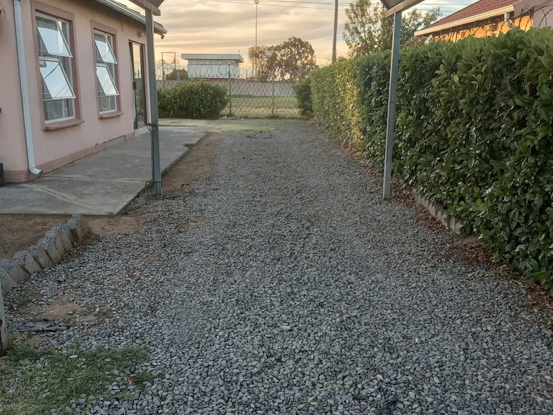 Rear exterior view featuring a gravel driveway, covered carport structure, concrete walkway, and established perimeter hedging alongside the house facade.