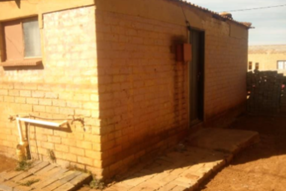Rear view of a basic brick structure featuring exposed brickwork, a corrugated metal roof, and external plumbing connections on a dirt lot.