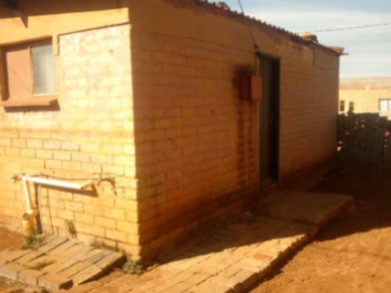 Rear view of a basic brick structure featuring exposed brickwork, a corrugated metal roof, and external plumbing connections on a dirt lot.