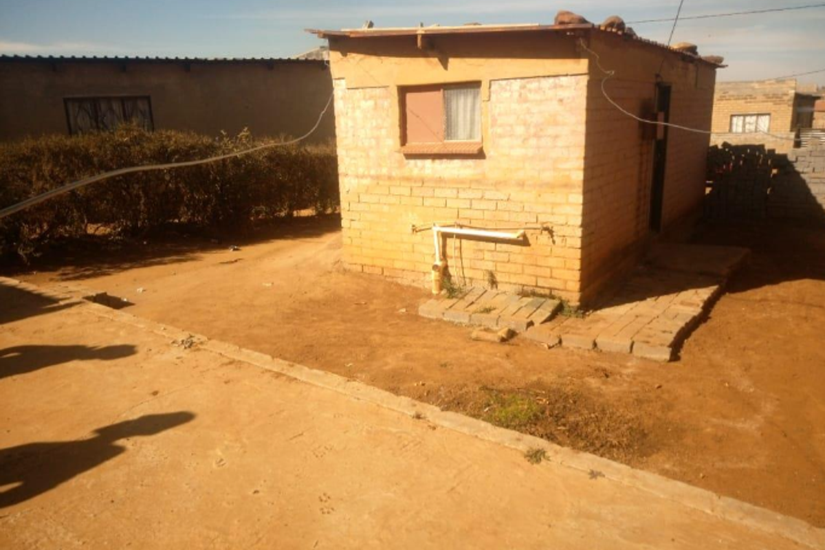 A detached brick outbuilding featuring exposed external plumbing and a simple roof structure, situated on a dirt yard.