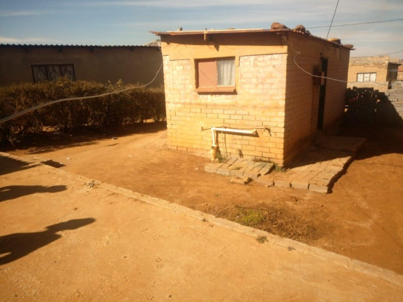 A detached brick outbuilding featuring exposed external plumbing and a simple roof structure, situated on a dirt yard.