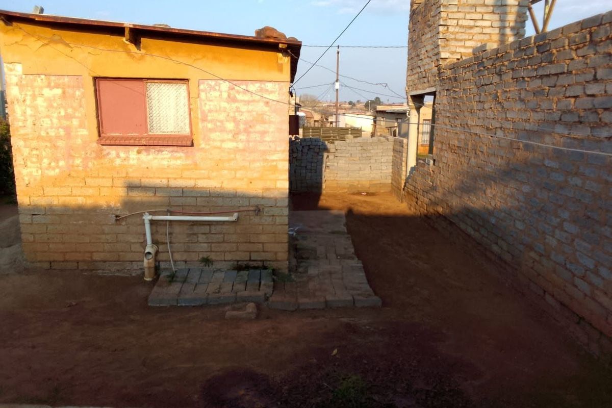 Rear exterior view of the property showing unfinished brick structures, exposed plumbing, and an unpaved yard area.