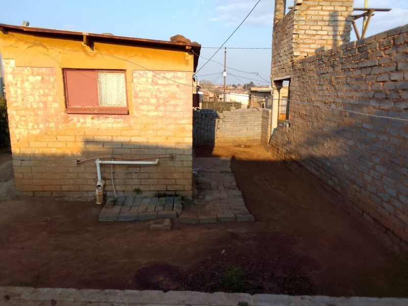 Rear exterior view of the property showing unfinished brick structures, exposed plumbing, and an unpaved yard area.