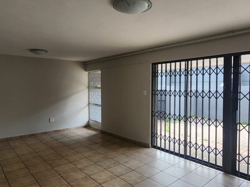 An empty, tiled living area featuring a sliding glass door with a security gate, providing direct access to an outdoor paved space.
