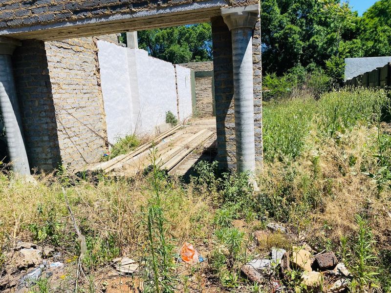 An unfinished residential structure featuring exposed brick masonry and concrete columns, situated on a plot with significant overgrown vegetation and debris.