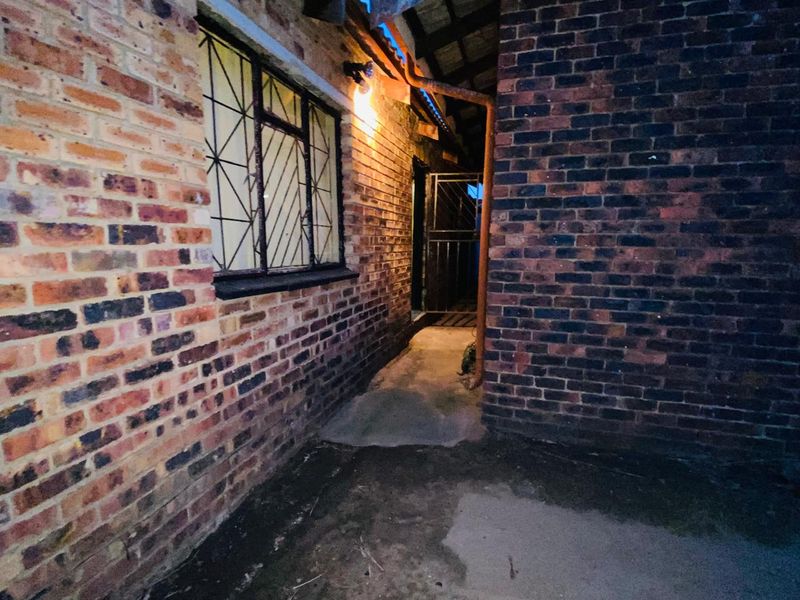 A narrow exterior rear passage featuring exposed brick walls, security bars on the window, and basic concrete paving under evening lighting.