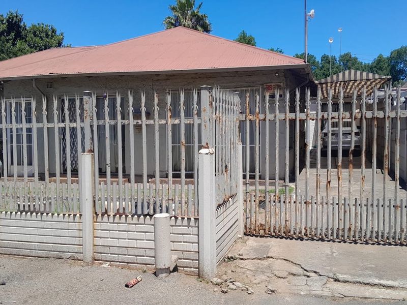 Front elevation of a single-story house featuring a metal security fence, gated driveway, and corrugated metal roof.